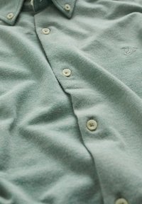Ljust grön skjorta med knappar i vitt och subtilt broderad logotyp på bröstet, visad på nära håll med tygveck.
