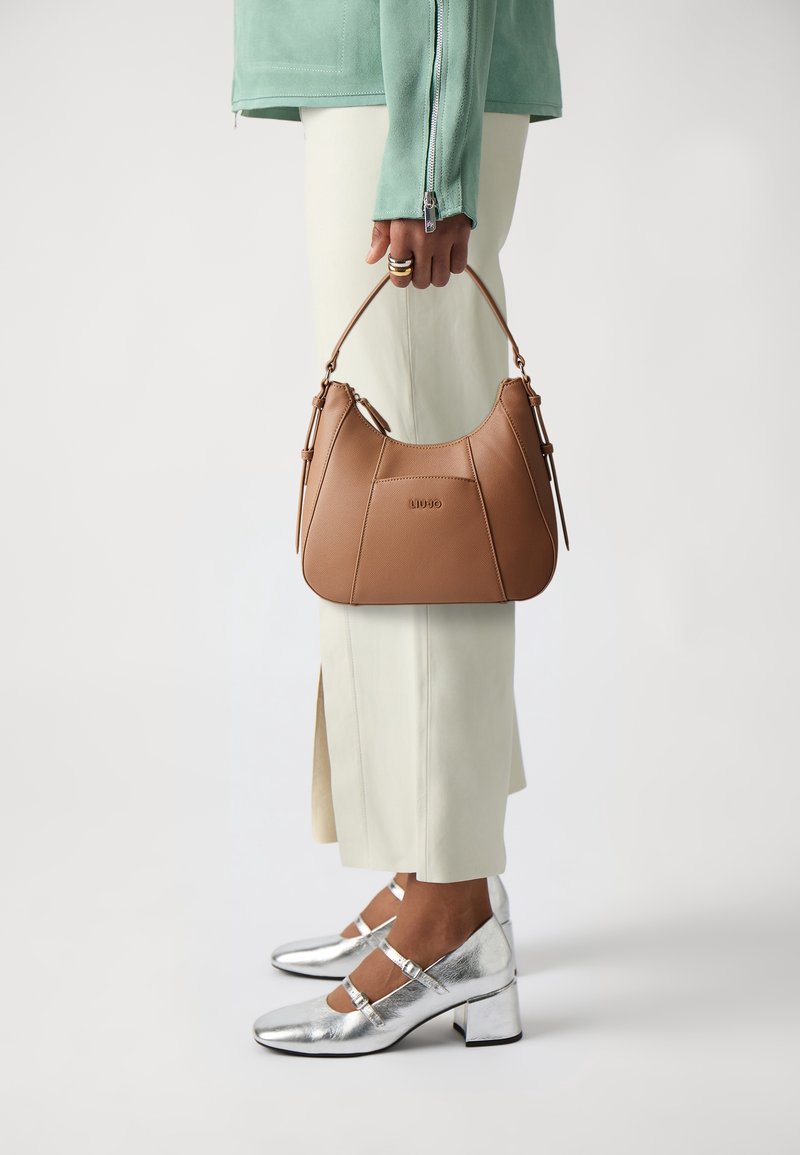 Personne portant un pantalon large crème et des talons Mary Jane argentés tenant un sac à main en cuir marron clair avec une anse supérieure et une fermeture éclair.