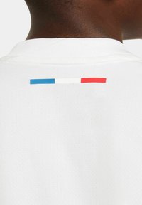 Chemise en tissu blanc avec un détail de rayures bleues, blanches et rouges au niveau du cou au dos. La texture est lisse avec un design simple.