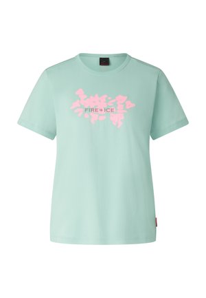 Personne portant de grandes lunettes de soleil à visière réfléchissante, t-shirt vert menthe, pantalon blanc ample, baskets bleu sarcelle et bandeau noir, debout les mains dans les poches.