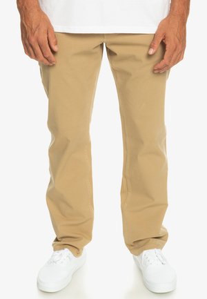 Light beige cotton pants with a straight-leg design, featuring back pockets and a smooth texture, paired with white sneakers.