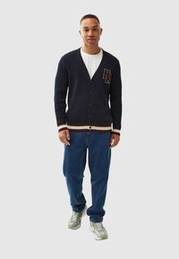 Marinblå ribbad cardigan med v-ringning, knäppning framtill och vinröda och krämfärgade randiga detaljer vid ärmar och nederkant. Har en logopatch på bröstet.