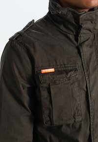 Mørkegrøn, bomuldsjakke med høj krave, lynlåse, lommer med knapper og orange logoetiket på brystet. Tekstureret og funktionelt design.