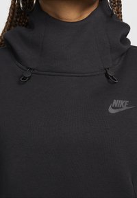 Crna dukserica s visokim ovratnikom, podesivim vezicom i suptilnim Nike logotipom. Glatka tekstura i minimalistički dizajn.