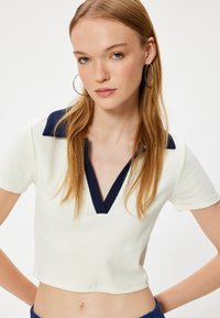 Korte mouw croptop in crèmewit met een marineblauwe kraag, met een V-hals ontwerp. De stof is geribbeld, wat een gestructureerde uitstraling geeft.