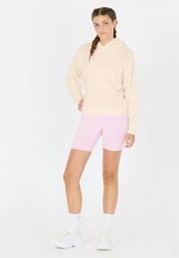 Beige Kapuzenpullover mit entspannter Passform, kombiniert mit rosa Radshorts. Das Modell trägt weiße Sneaker und knöchellange weiße Socken.