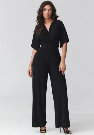 Kobieta z długimi falowanymi włosami, ubrana w czarny kombinezon z dekoltem w serek, szerokimi nogawkami i krótkimi rękawami, stojąca na jednolitym jasnym tle.