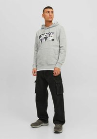 Sudadera gris con un estampado gráfico en negro y morado, con un bolsillo frontal, combinada con pantalones cargo negros y zapatillas grises. Diseño simple y casual.