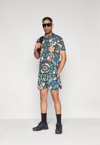 Σετ με κοντομάνικο πουκάμισο και σορτς με σκούρο λουλουδάτο μοτίβο που περιλαμβάνει πράσινο, μπλε, ροδάκινο και λευκό. Φοριέται με μαύρα παπούτσια και γυαλιά ηλίου.