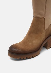 Bottine en daim marron avec un bout arrondi et lisse, un talon épais, et un panneau contrastant couleur beige. Semelle texturée pour une meilleure adhérence.