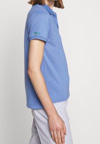 Ljusblå polotröja med krage, korta uppvikta ärmar och grön "Polo" brodyr på vänster arm. Tillverkad av en strukturerad tyg.