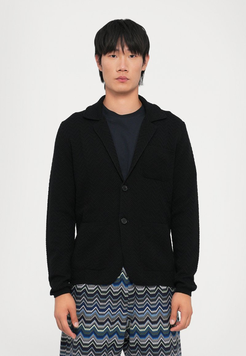 Melnā tekstūrā blakus ar šāla apkakli, vienu pogu aizdares un divām priekšējām kabatām, kombinēta ar rakstainām zilām un zaļām biksēm.