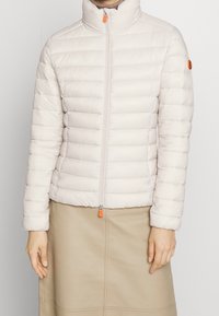 Light beige puffer jacket with quilted design, high collar, front zipper with orange pull tab, and long sleeves.