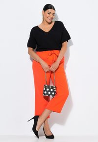 Top nero in maglia con scollo a V, pantaloni arancioni brillanti a vita alta con dettaglio a fiocco, décolleté neri in suede e una piccola borsa a pois bianco e nero.