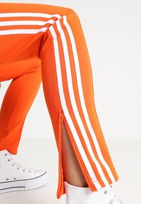 Jalka koukussa, yllään oranssit housut, joissa on valkoiset raidat ja sivuvetoketju, yhdistettynä valkoisiin korkeisiin tennareihin valkoista taustaa vasten.