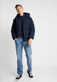 Chaqueta acolchada azul marino con capucha, cierre frontal y puños elásticos, combinada con un suéter azul marino y jeans de color azul claro. El modelo lleva zapatillas negras.