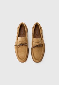 Mocassins en suède marron avec un design courbé, présentant un rabat et un détail de lacets décoratifs. Intérieur en cuir lisse et semelle en caoutchouc.