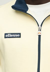 Veste zippée jaune pâle avec un col bleu marine. Présente un logo brodé sur la poitrine et un tirette de fermeture éclair avec un accent coloré.