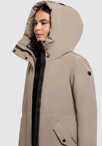 Beige geïsoleerde jas met grote capuchon, rits- en drukknoopsluitingen, twee zijzakken en een gladde buitenkant. Zwarte accenten op de rits.