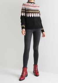 Czarny sweter w sploty z wzorzystym karczkiem w odcieniach różu, czerwieni, żółci i bieli. Połączony z ciemnymi obcisłymi dżinsami i czerwonymi butami na obcasie z ćwiekami.