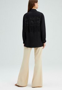 Zwarte blouse met lange mouwen, versierd met bloemenkant en franjes op de rug, gecombineerd met crèmekleurige wijde broek. Gladde textuur, met kragen.