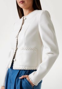 Witte leren jas met een getailleerd ontwerp, decoratieve stiksels langs de randen, twee zakken en een knoopsluiting aan de voorkant.