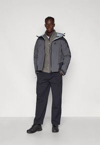 Grijze geïsoleerde jas met capuchon over een grijze trui, gecombineerd met donkere broek en zwarte schoenen. Eenvoudig ontwerp met contrasterende kleuren.