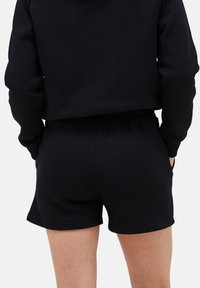 Schwarze Sweatshorts mit elastischem Bund. Glatte Textur, figurbetontes Design und seitliche Taschen. Passender schwarzer Pullover ist teilweise sichtbar.