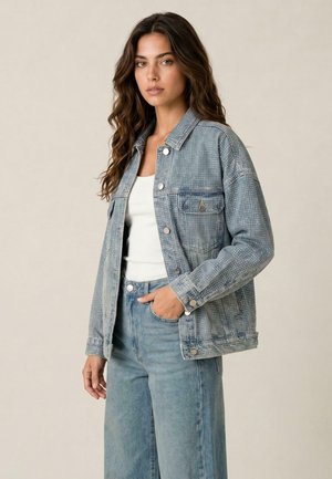 Donna con lunghi capelli castani che indossa una giacca di jeans azzurro chiaro con puntini bianchi, una maglietta bianca e jeans blu, in piedi con una mano in tasca.