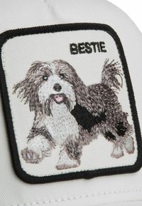 Écusson brodé d’un chien heureux à poils longs, avec un pelage noir et blanc, langue tirée, au-dessus du mot « BESTIE » sur un tissu blanc bordé de noir.