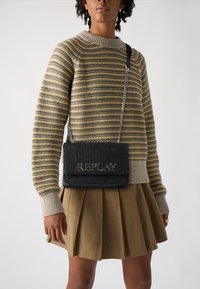 Borsa a spalla nera con texture, dotata di tracolla in catena argentata e logo "REPLAY", abbinata a un maglione a righe e una gonna plissettata beige.