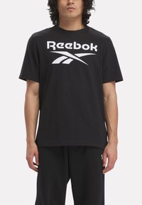Melnā kokvilnas t-krekliņa ar lielu baltu Reebok logo un stilizētu grafiku. Īsās piedurknes un klasiska apaļa kakla izgriezne.