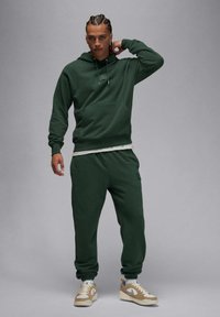 Felpa con cappuccio verde scuro con tasca frontale, abbinata a pantaloni jogging coordinati. Tessuto strutturato, vestibilità morbida e sneakers con accenti beige.