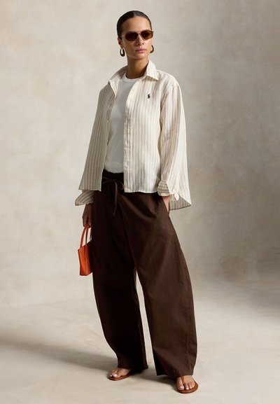 Mujer con camisa a rayas beige, top blanco, pantalones marrones de pierna ancha, sandalias, gafas de sol, pendientes de aro, sosteniendo un pequeño bolso naranja.