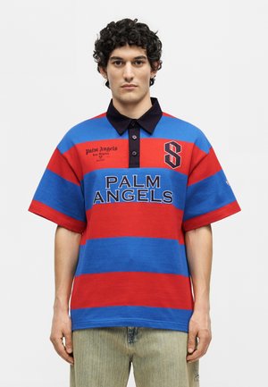 Άνδρας με σγουρά μαλλιά που φορά μπλε και κόκκινο ριγέ πόλο μπλουζάκι με το κείμενο "Palm Angels" και τον αριθμό 8 στο στήθος, στέκεται μπροστά σε απλό φόντο.