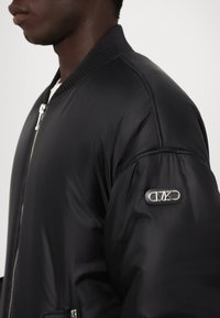 Doudoune noire avec col en rib, fermeture éclair et poches latérales. Présente un patch logo sur la manche gauche et une texture de tissu lisse et brillante.