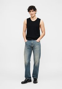 Νέος άνδρας με σγουρά μαλλιά που φοράει μαύρο αμάνικο τοπ, ξεβαμμένο μπλε τζιν και μαύρα παπούτσια, στέκεται με τα χέρια στις τσέπες.