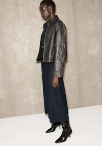 Chaqueta de cuero negra con un diseño ajustado, llevada sobre un vestido azul oscuro, combinada con botines de tacón negro puntiagudos, contra un fondo neutro.