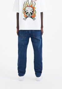 Camiseta blanca con un gráfico de calavera colorido y un diseño de llamas en la parte posterior; pantalones vaqueros de mezclilla azul con un ajuste relajado y dos bolsillos traseros.