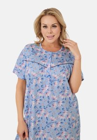 Blondynka w niebieskiej sukience w kwiaty z krótkim rękawem i guzikami, uśmiechająca się i dotykająca swoich włosów na jednolitym tle.