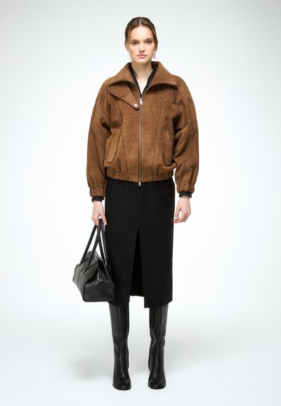 Žena v hnedej búndě na zips, čiernej midi sukni s predným rozparkom, čiernych vysokých čižmách prevyšujúcich kolená, držiaca čiernu koženú kabelku.