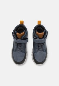 Zapatos azul marino con parte superior de cuero texturizado, suela de goma gris y un tirador amarillo. Presentan un diseño con cordones en la parte frontal y una correa para el tobillo.