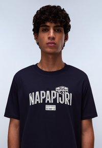 Námorně modré bavlněné tričko s bílým textem "NAPAPIJRI" a logem na horní levé straně. Krátké rukávy a kulatý výstřih. Hladká textura.