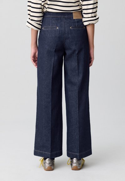 Jeans de mezclilla oscuros de pierna ancha con bolsillos traseros y detalles en dorado, combinados con zapatos metálicos y una camiseta a rayas.