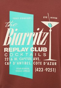 Καφέ βαμβακερό t-shirt με γραφικό σχέδιο σε τουρκουάζ και λευκό, κείμενο για το "the Biarritz Replay Club" και εικονογράφηση ποτηριού μαρτίνι.