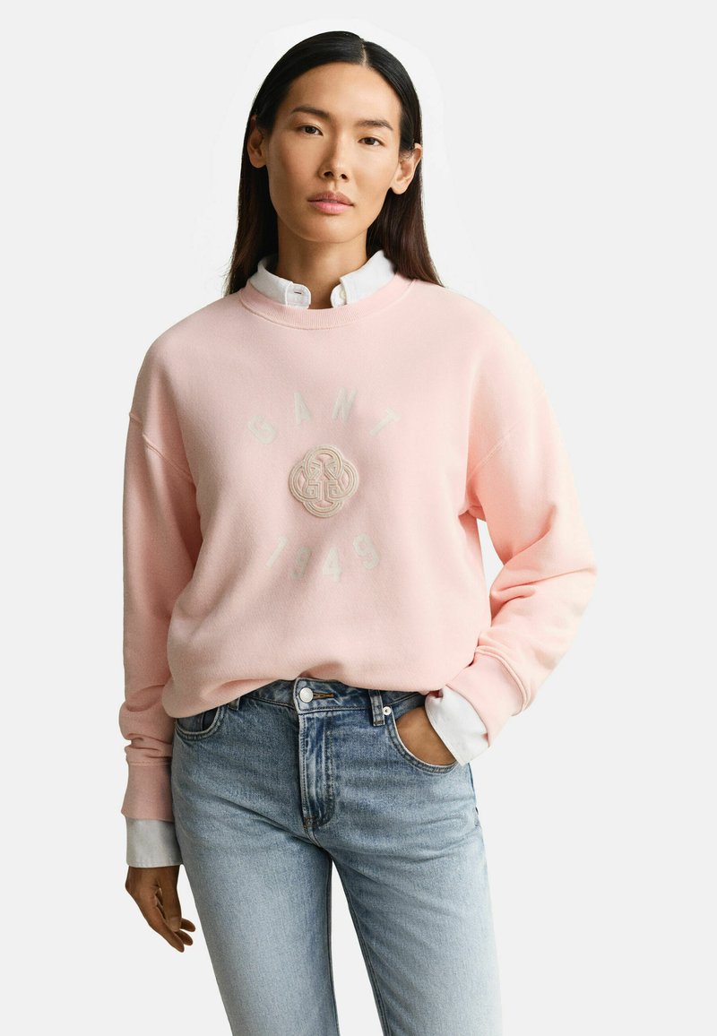 Mujer con cabello largo y oscuro que lleva una sudadera rosa claro de GANT sobre una camisa blanca abotonada, jeans azul claro y con una mano en el bolsillo.