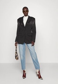 Moteris dėvi juodą švarką su kutais, baltą aukštu kaklu megztinį, šviesiai mėlynas džinsines kelnes, rudus aukštakulnius su raštais ir laiko mažą raštuotą rankinę.