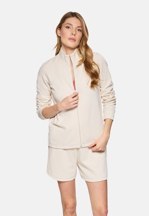 Mujer con cabello largo que lleva una chaqueta beige con cremallera y pantalones cortos a juego, parcialmente desabrochando la chaqueta para revelar una camiseta roja debajo.