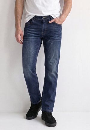 Homme portant un jean bleu foncé avec des détails délavés, un t-shirt blanc et des baskets noires, debout contre un mur blanc.