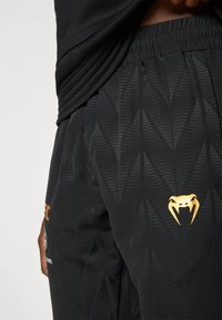 Μαύρο αθλητικό σορτς με υφή που απεικονίζει σχεδιασμούς σε μορφή κυμάτων. Στη μία πλευρά υπάρχει ένα χρυσό λογότυπο φιδιού.